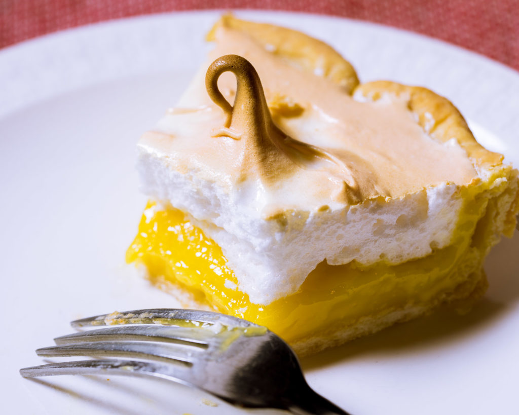 A half eaten slice of lemon meringue pie. A fork sits in front of the slice.